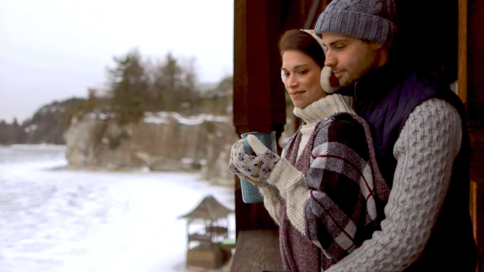 Couple wearing warm winter clothing. They are standing on a balcony overlooking a lake drinking
