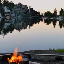 Sunsetting in the background. There is a campfire with benches surrounding it in front of a lake. In the background there is a large hotel