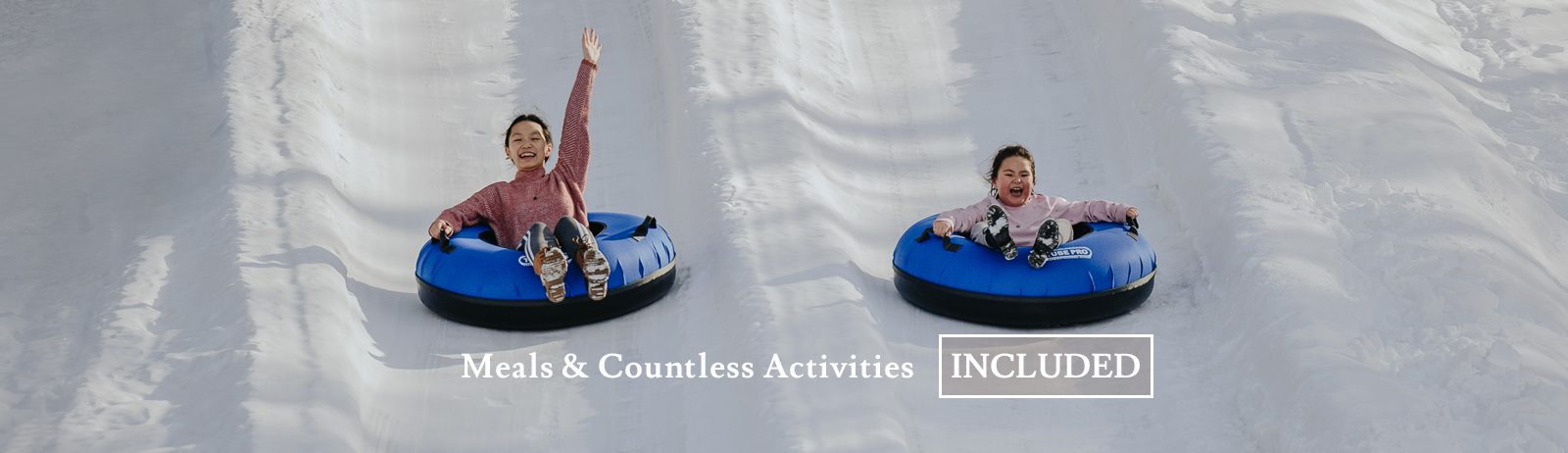 Image of two children smiling, sliding down a snow covered hill on snow tubes